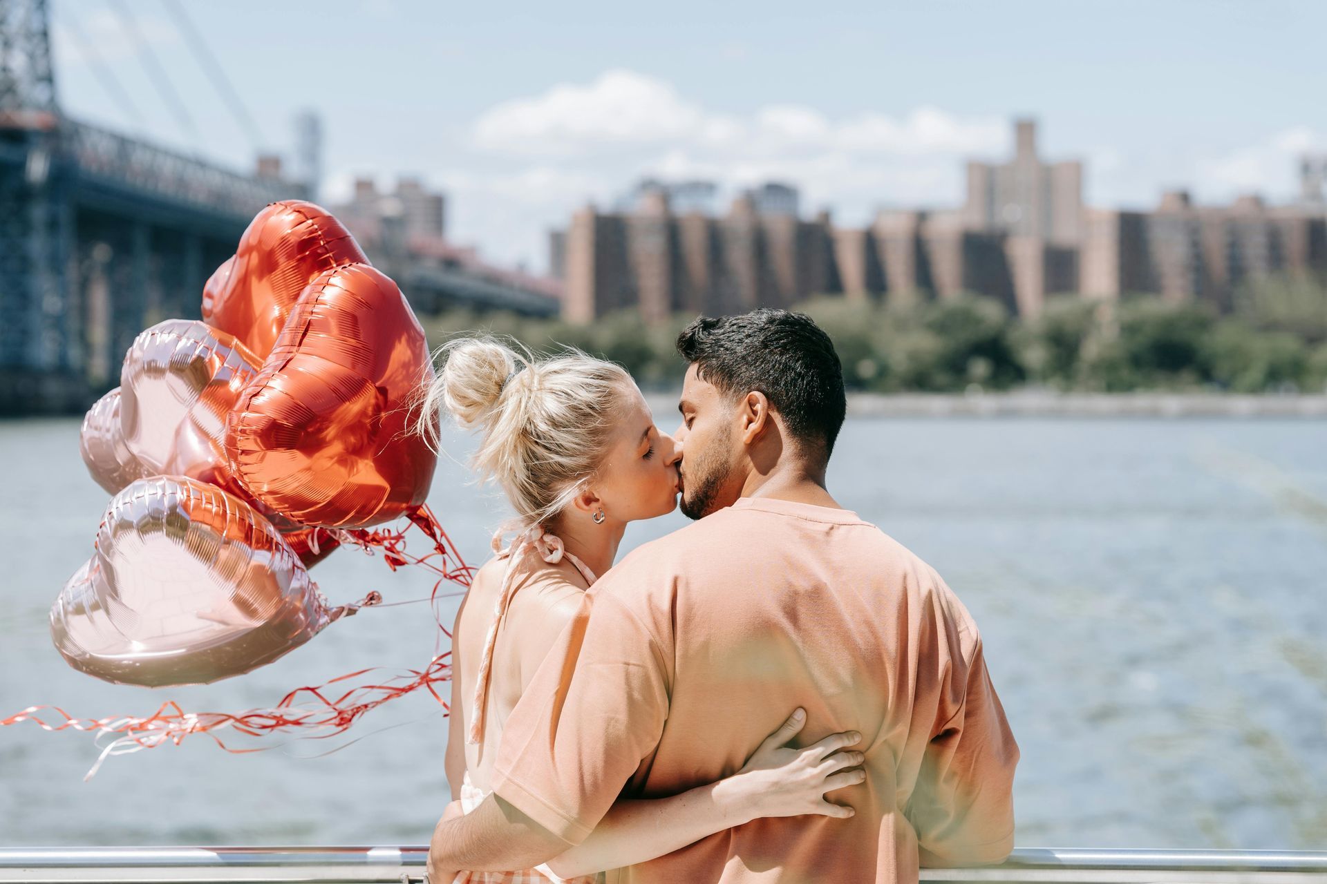 Couple kissing near water, holding heart balloons in New York City.