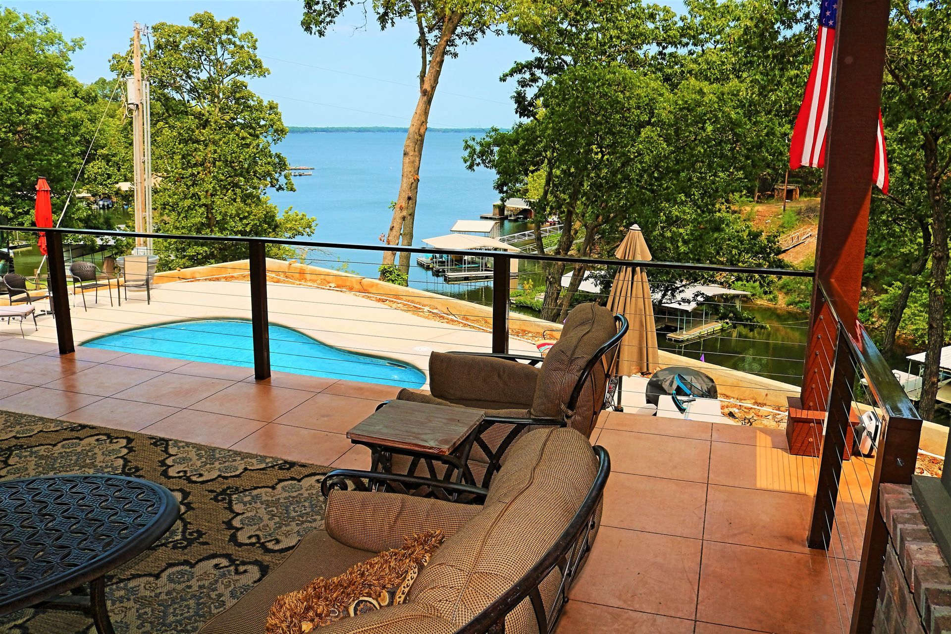 Deck overlooking a lake with a pool, trees, and patio furniture. American flag displayed.