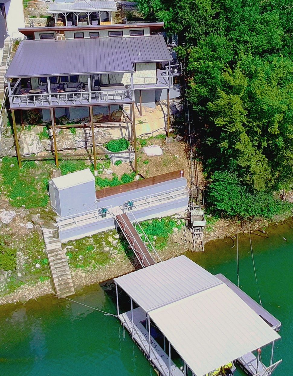 Waterfront house on stilts with a dock and boat. Green water and trees surround it.