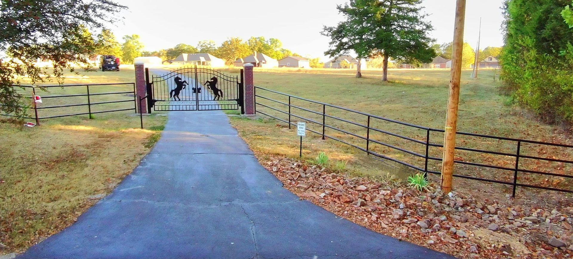 A driveway leading to a gated property with decorative ironwork; a field and trees in the background.