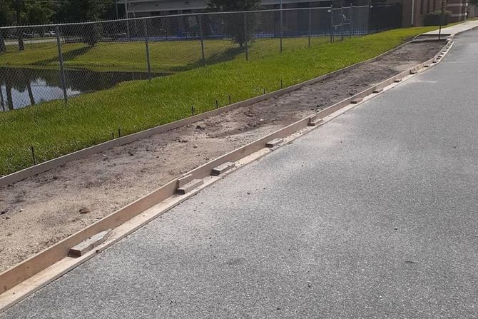 Roadside curb with gravel shoulder, grassy embankment, and chain-link fence beside pavement