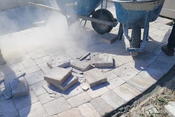 Construction workers pouring concrete onto a tiled patio beside a blue mixer, with stacked pavers nearby