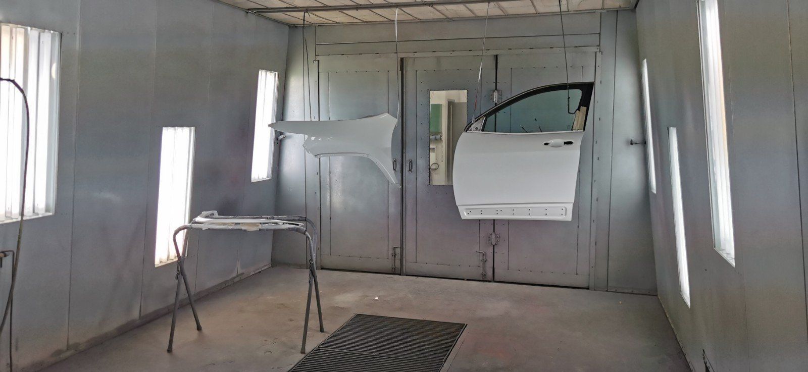 Une portière et un garde-boue de voiture blancs sont suspendus dans une cabine de peinture avec des lumières et une table.