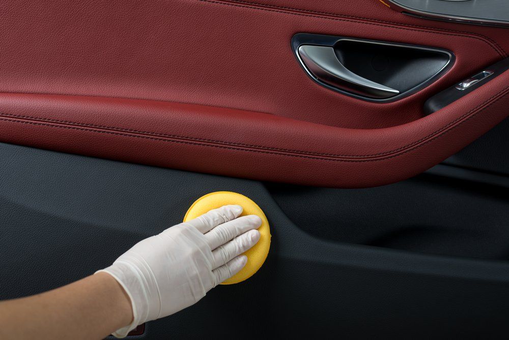 Une main gantée nettoie le panneau de porte noir d'une voiture avec une éponge jaune. On aperçoit les garnitures de porte en cuir rouge.