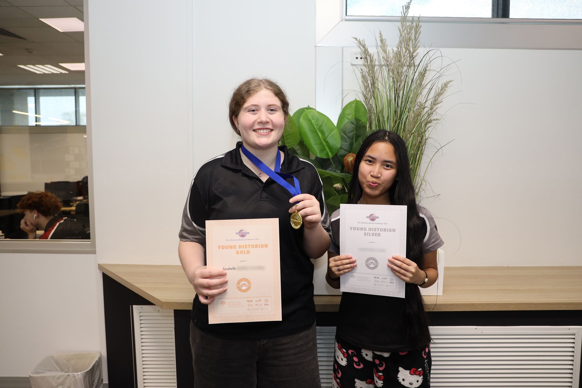 Two people holding certificates and a medal indoors, smiling. One is sticking out their tongue.