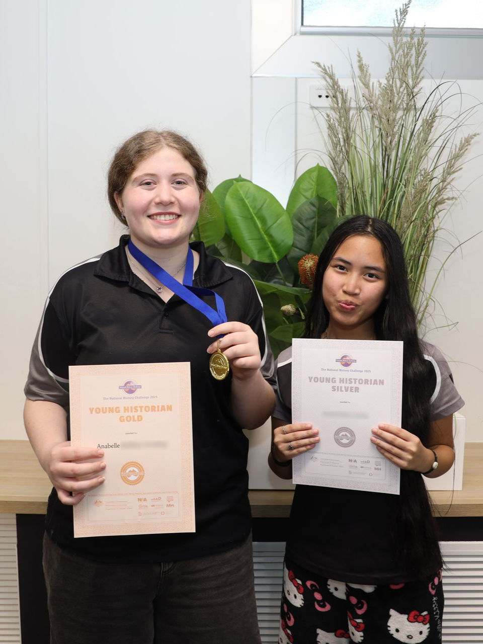 Two people holding certificates and a medal, smiling in front of a plant.
