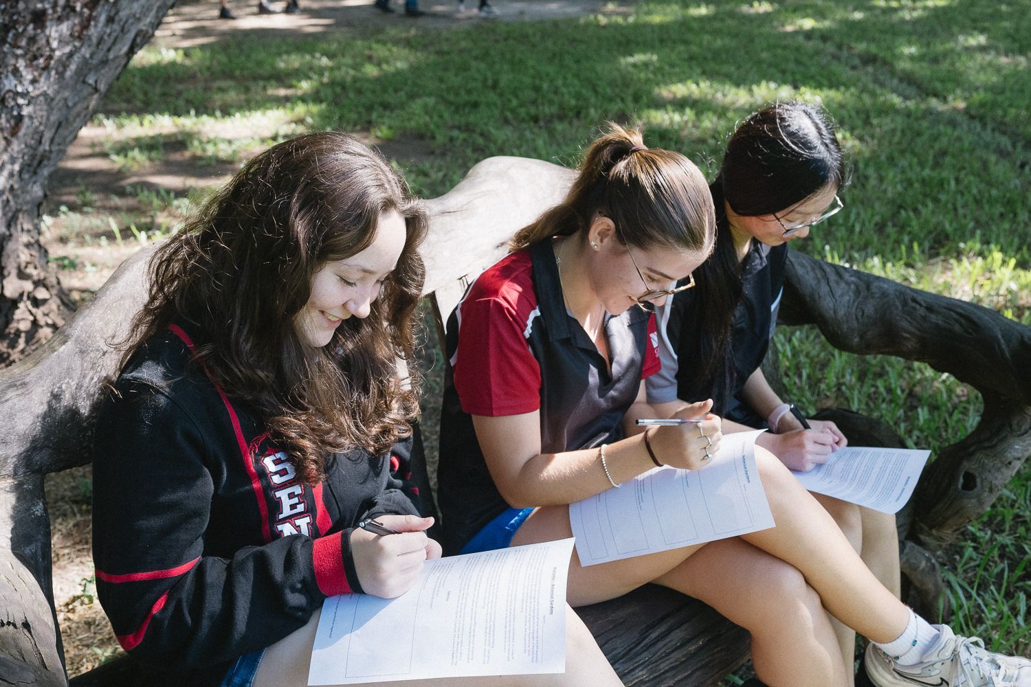 Three students write on paper while sitting on a log outdoors.