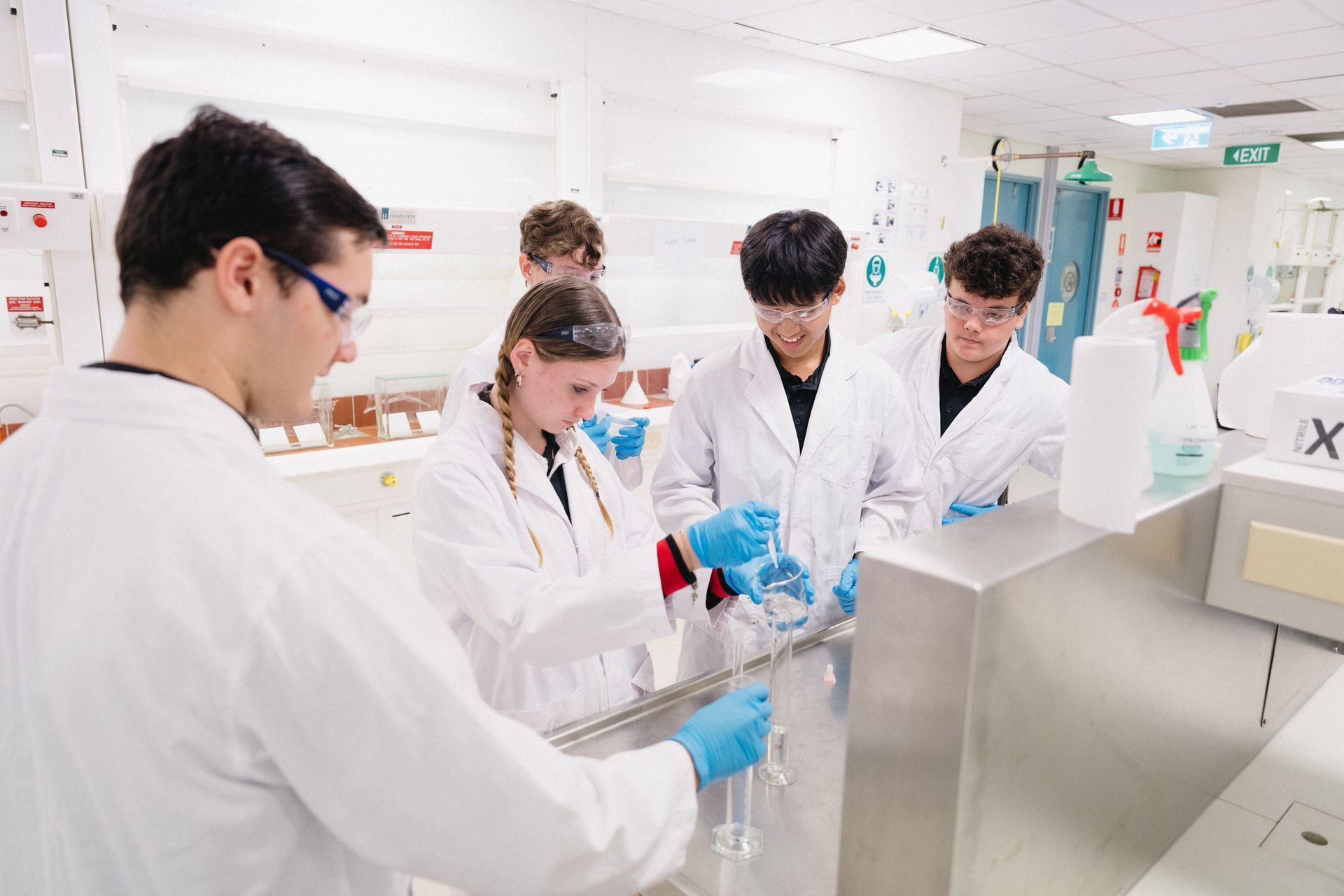 Students in lab coats and safety glasses work in a science lab, guided by an instructor.