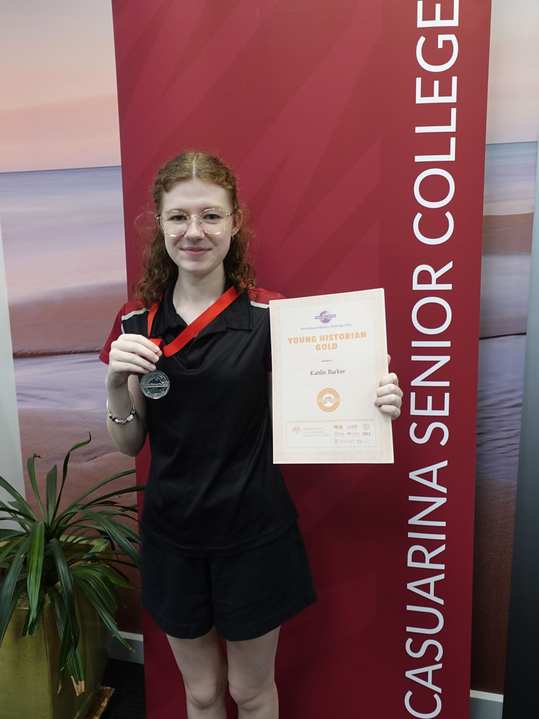 Teen holding a certificate and medal in front of a Casuarina Senior College banner.