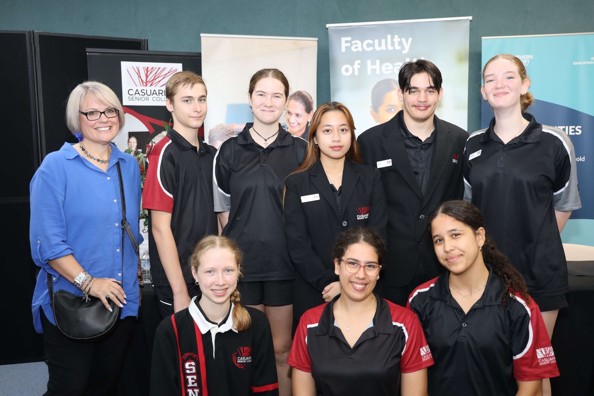 Group of people at a faculty event, smiling. Some wear black & red uniforms, others in dress clothes. Banners in background.