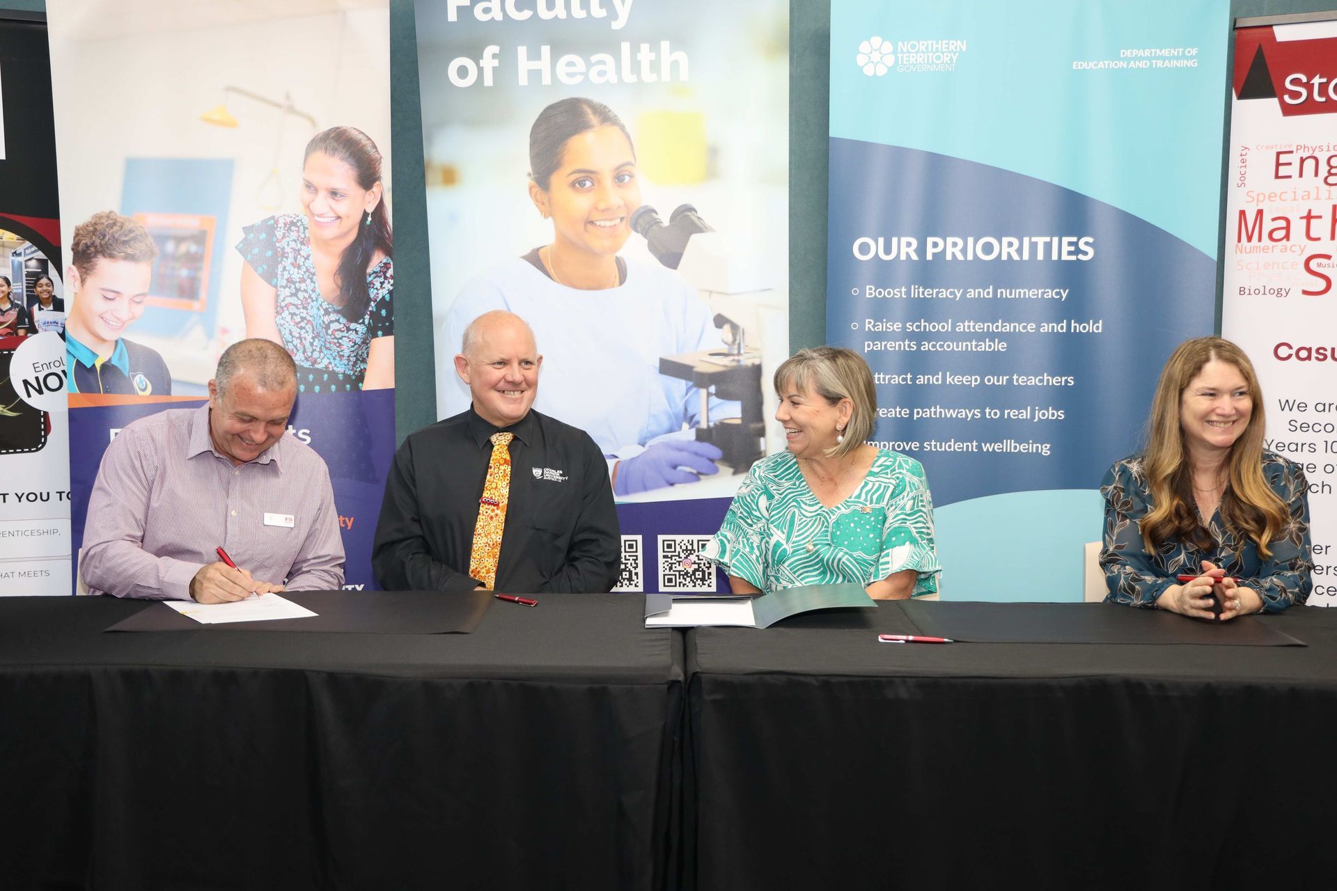 Four people seated at a table signing documents, behind banners for the Faculty of Health and STEM programs.