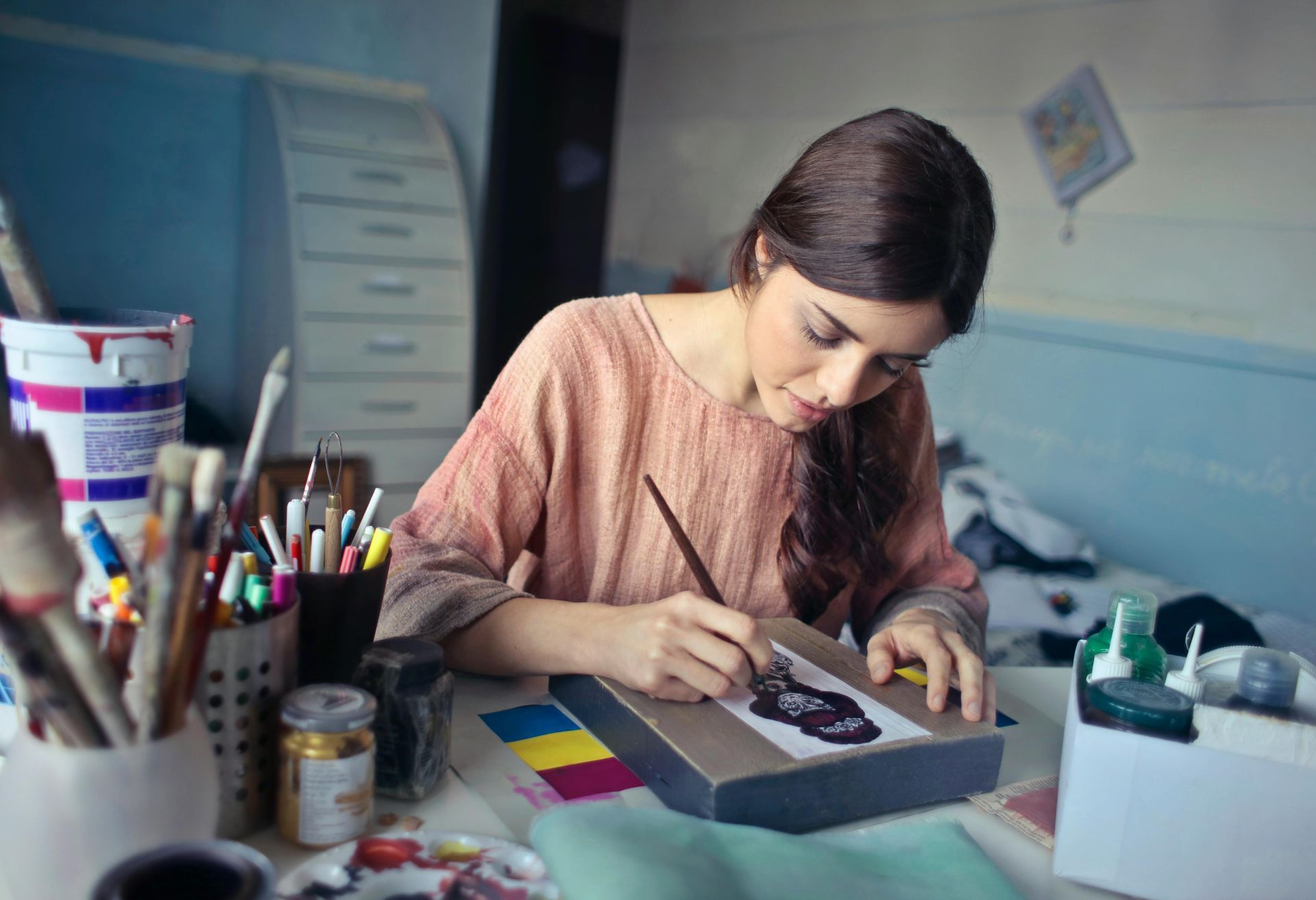 Woman painting at a desk, surrounded by art supplies, in a room with blue walls.