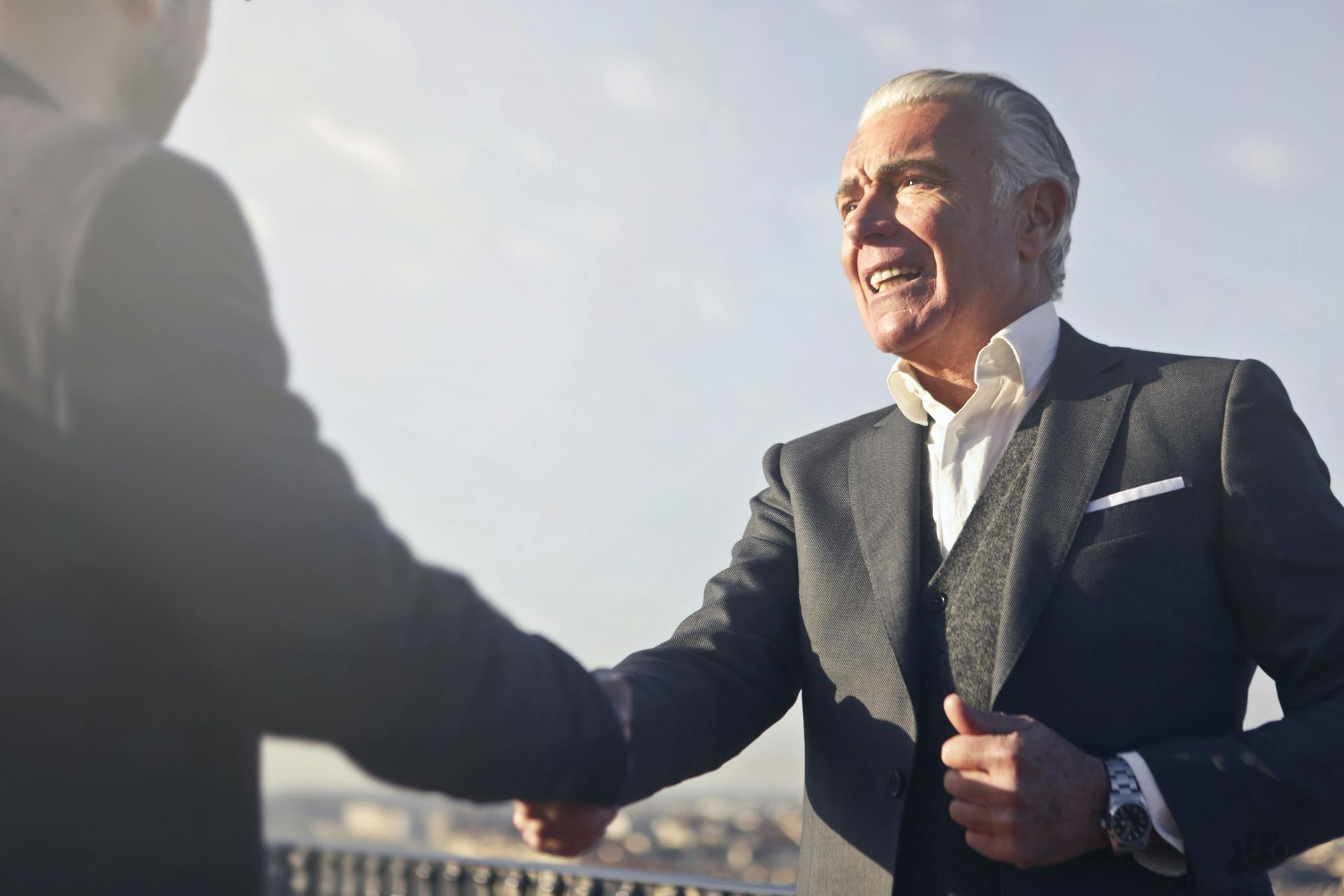 Two businesspeople shaking hands, smiling. One in dark suit, white shirt, and wristwatch. Outdoor setting.