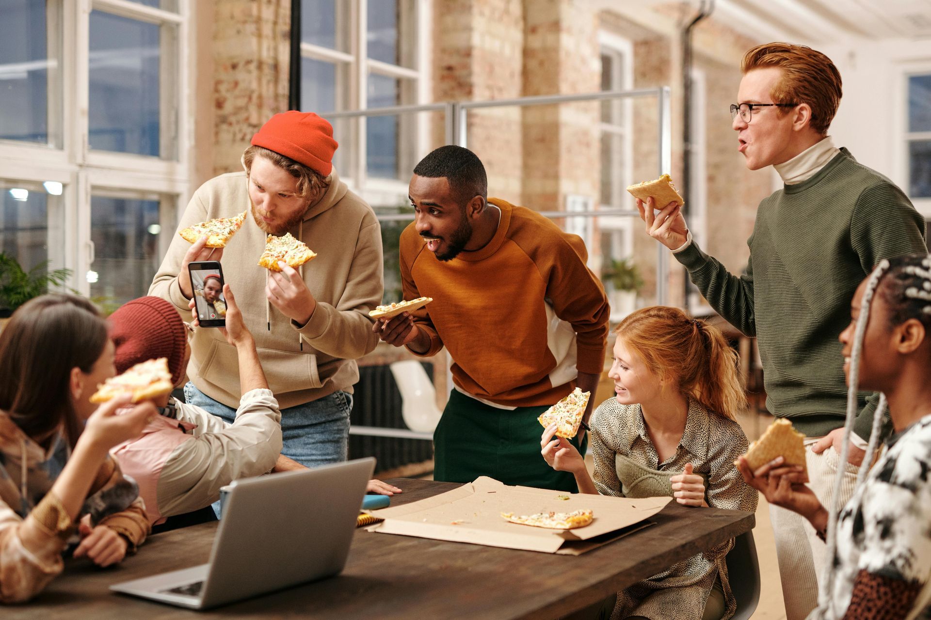 Group of people eating pizza around a table in an office setting; some are holding pizza slices and smiling.