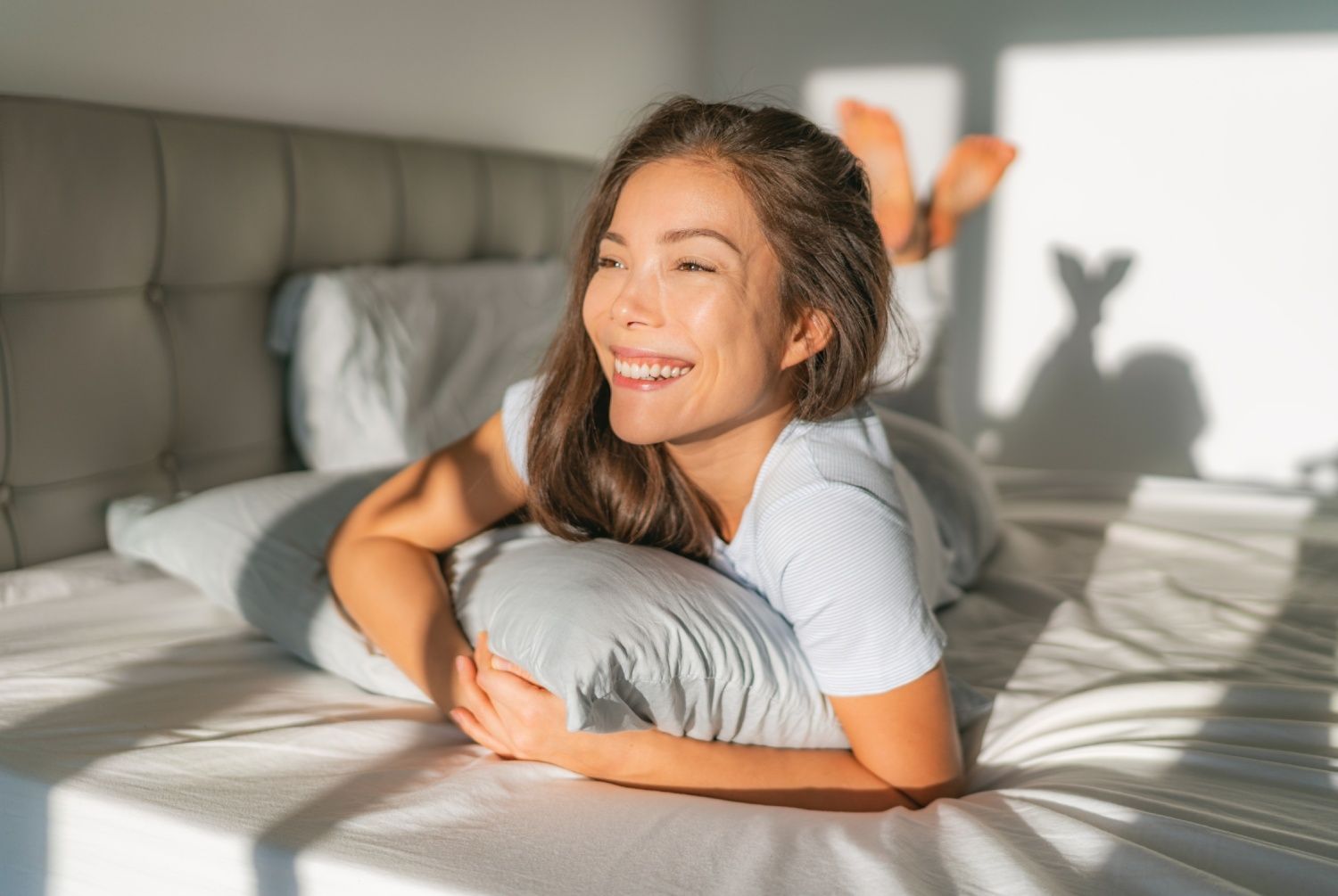 Woman smiling, lying on bed, propped on pillow, sunlight, shadow on wall.
