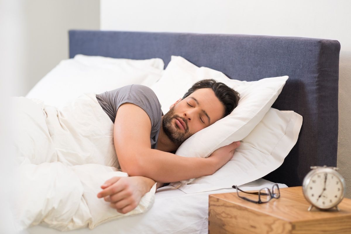 Man asleep in bed, white bedding, blue headboard, side table with glasses and alarm clock.