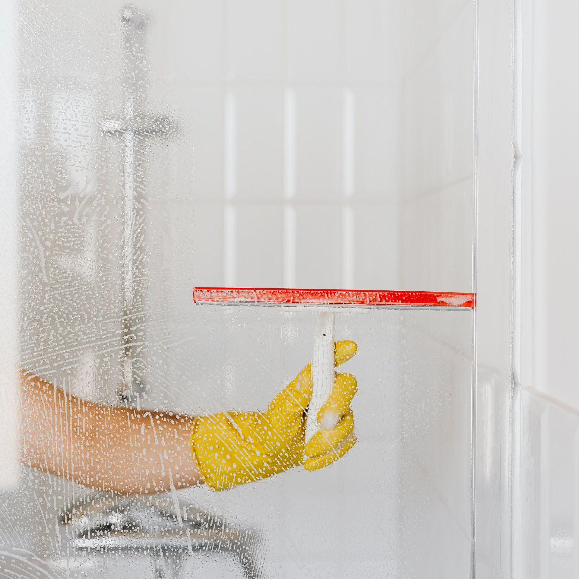A person wearing yellow gloves is cleaning a shower door with a squeegee.