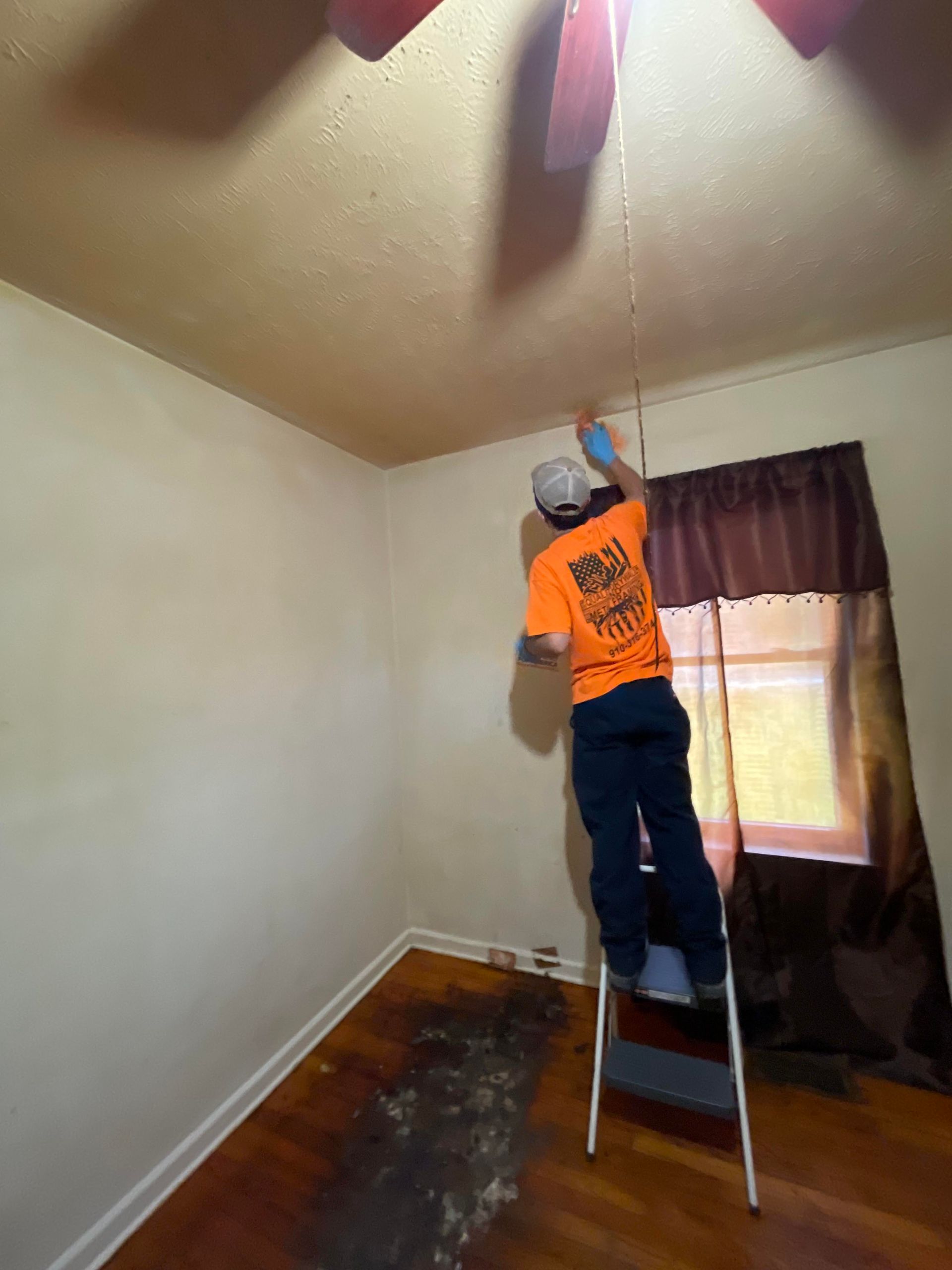 A man in an orange shirt is standing on a ladder in a room