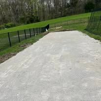 Gravel path bordered by black fences and green grass in a grassy field, trees in the background.