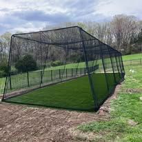 A black baseball batting cage on artificial turf in a grassy outdoor area.