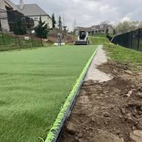Green artificial turf being installed next to a dirt path and a small construction vehicle in a residential area.
