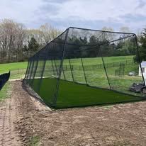 Black batting cage on green turf, set outdoors on dirt ground.