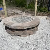 Stone fire pit filled with gravel, set on a gravel surface. Wooden posts in background.