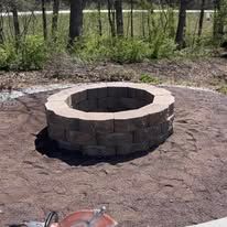 Stone fire pit on a bed of tan gravel, surrounded by dirt and trees.
