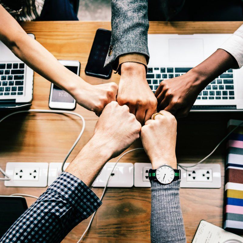 Fists bumping over a table with laptops and phones, symbolizing teamwork and collaboration.