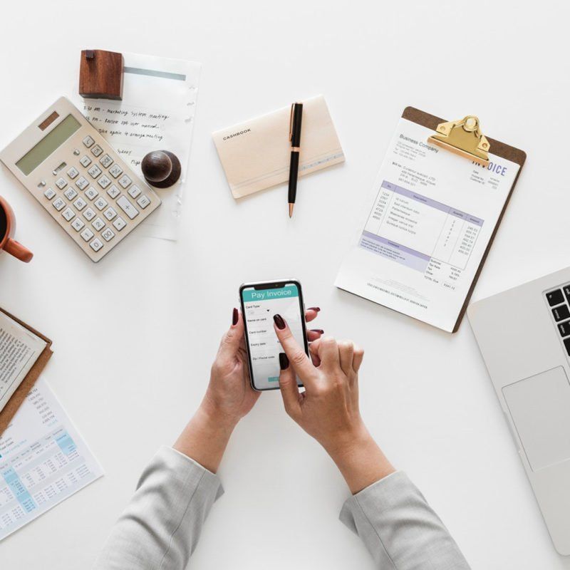 Hands using a smartphone on a white desk surrounded by documents, calculator, pen, and laptop.
