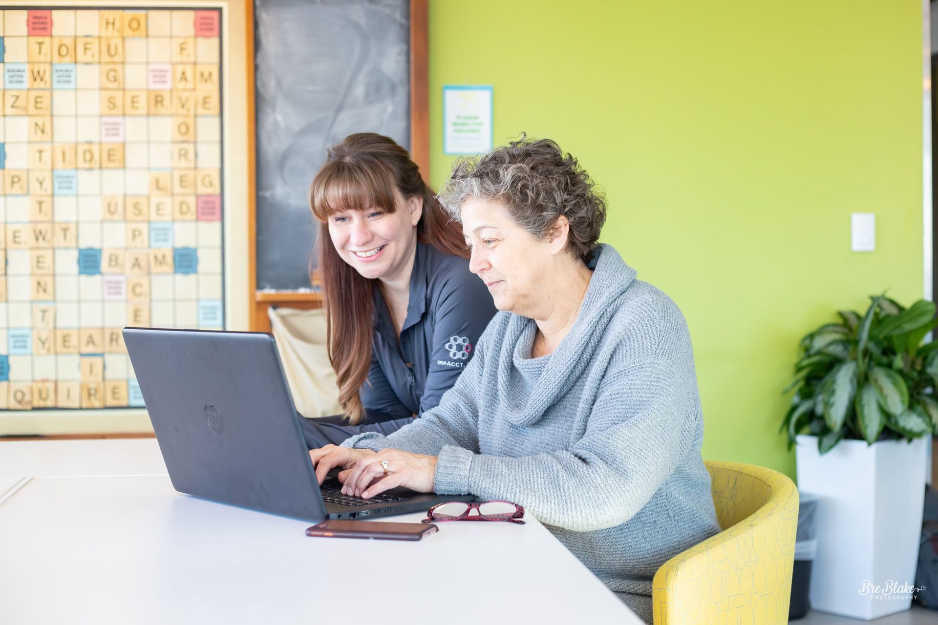 Woman assists another with a laptop, both smiling, in a room with a Scrabble board and green wall.