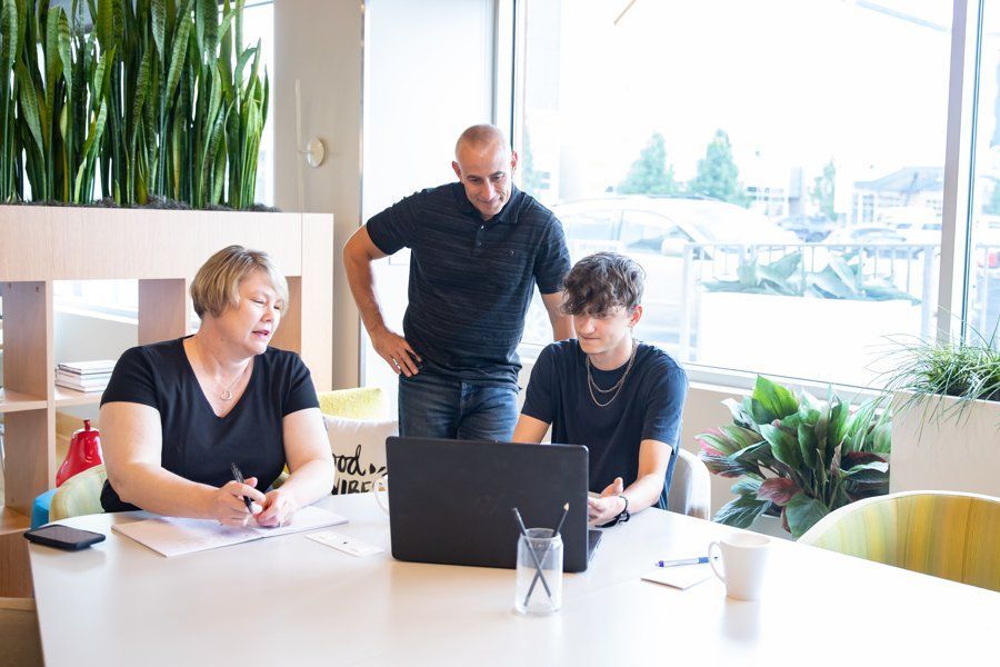 Three people reviewing a laptop at a table in a bright office.