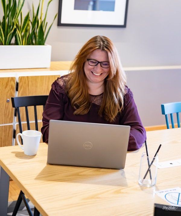 Woman smiling while using a laptop at a table, coffee mug, and a glass of straws nearby.