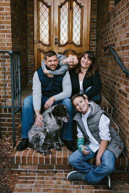 Family and dog pose on brick steps. A boy hugs his dad, another smiles. Woman and dog are also present.