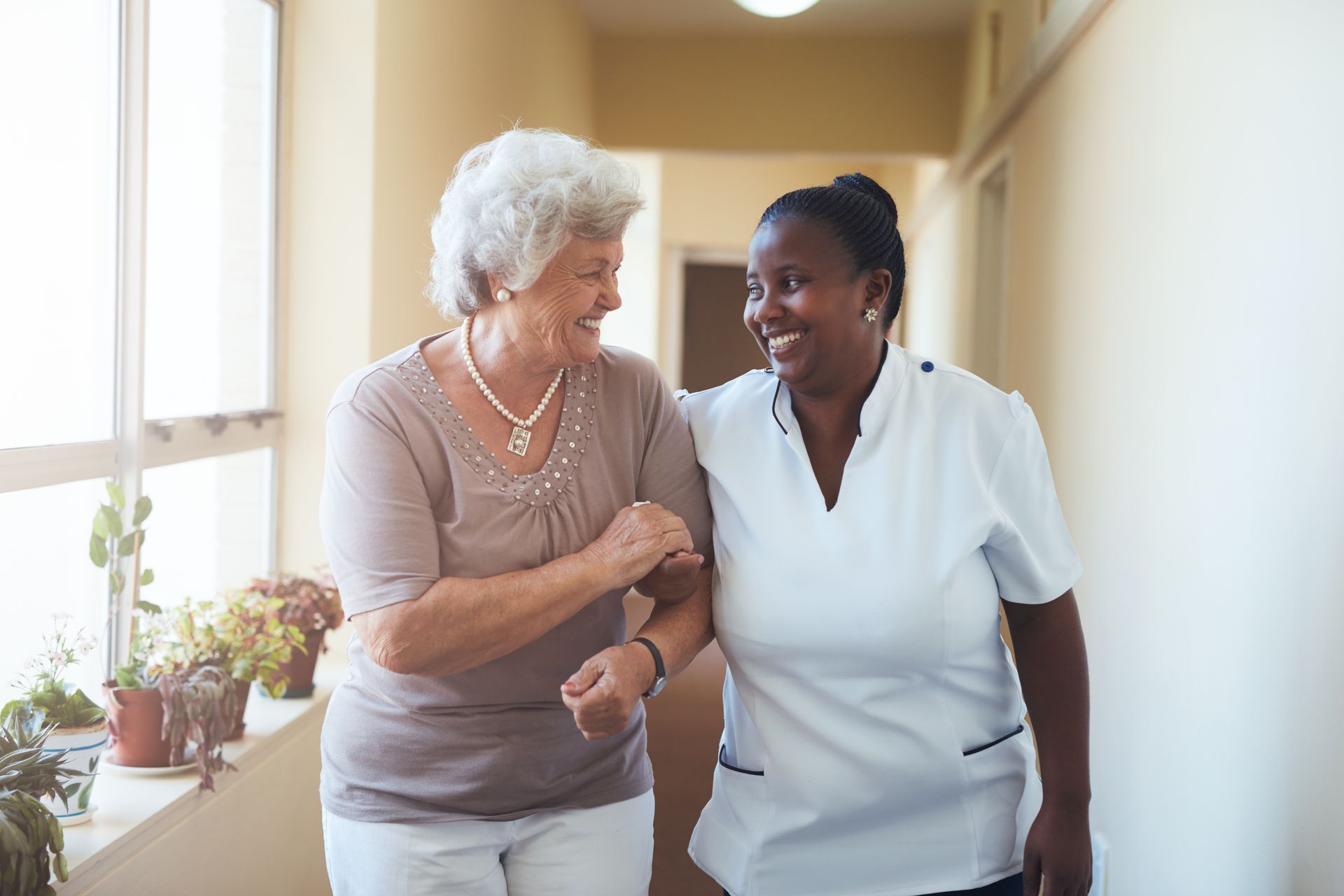 Woman assisted by a caregiver walking down a well-lit hallway. Both smile.