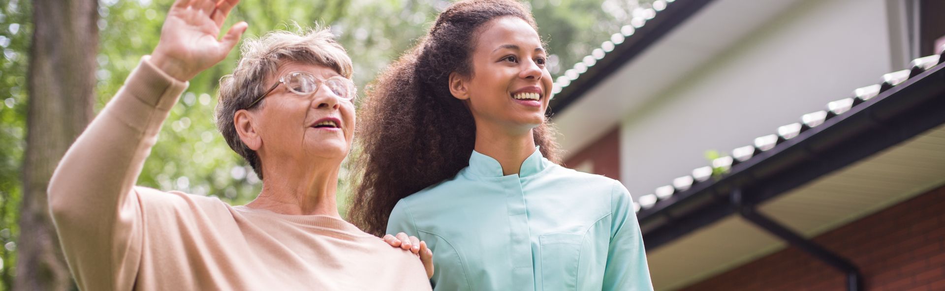 A woman caregiver is standing beside an old woman outside a house, resting a hand on her arm
