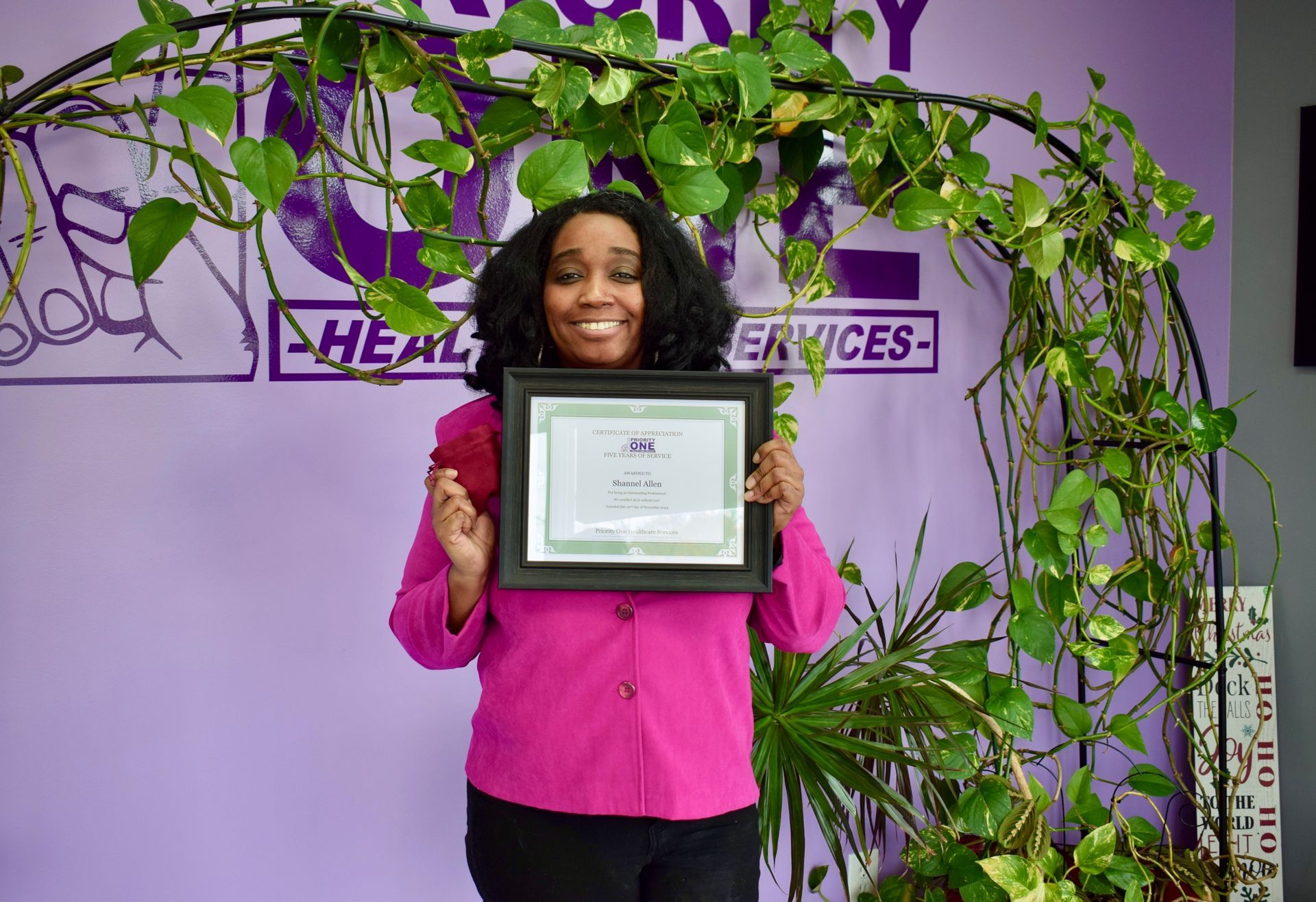 A Woman in A Pink Shirt Is Holding a Certificate in Front of A Purple Wall.