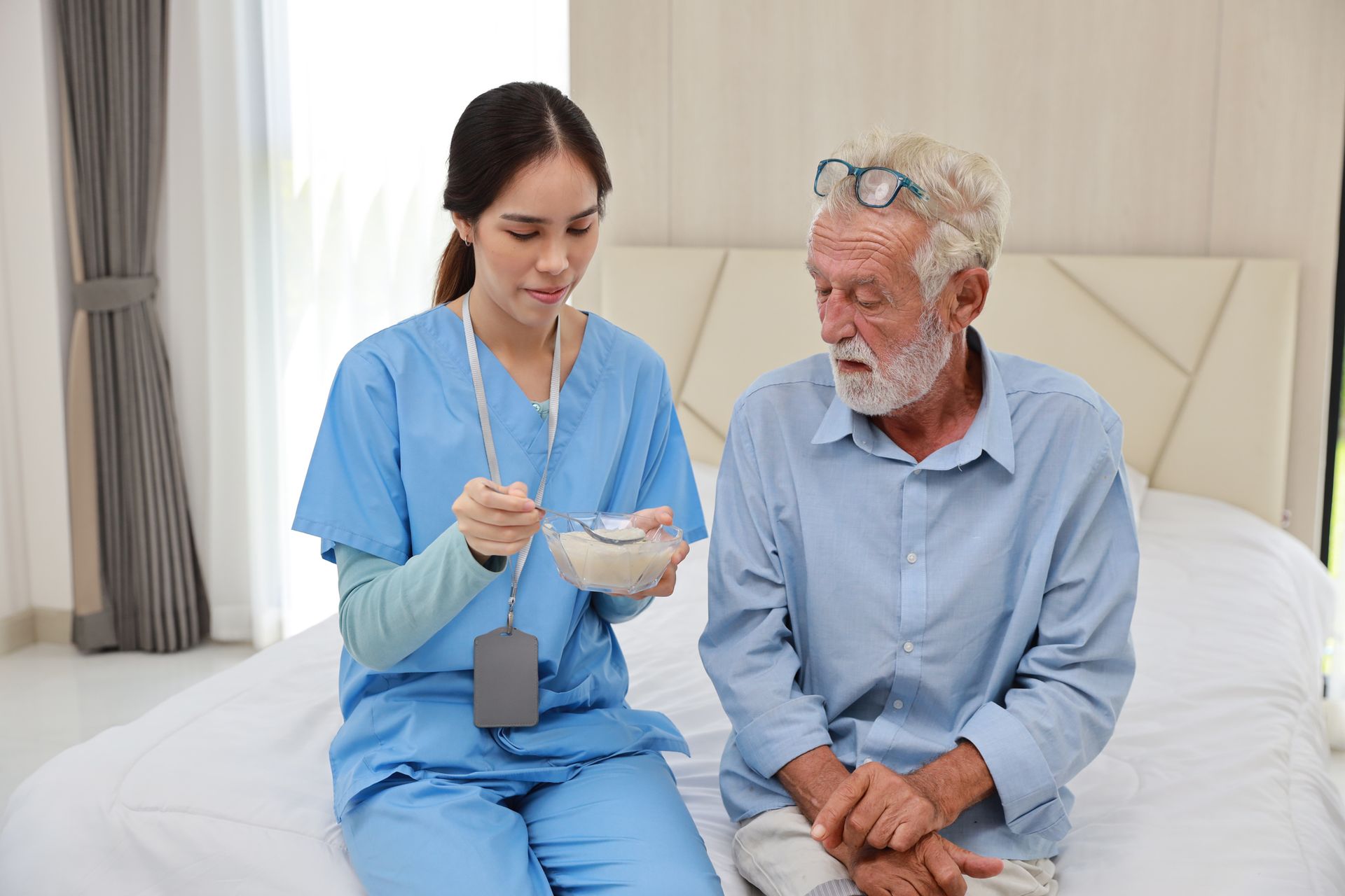 A caregiver feeding an old senior man in a bed with rice porridge soup for breakfast at his house