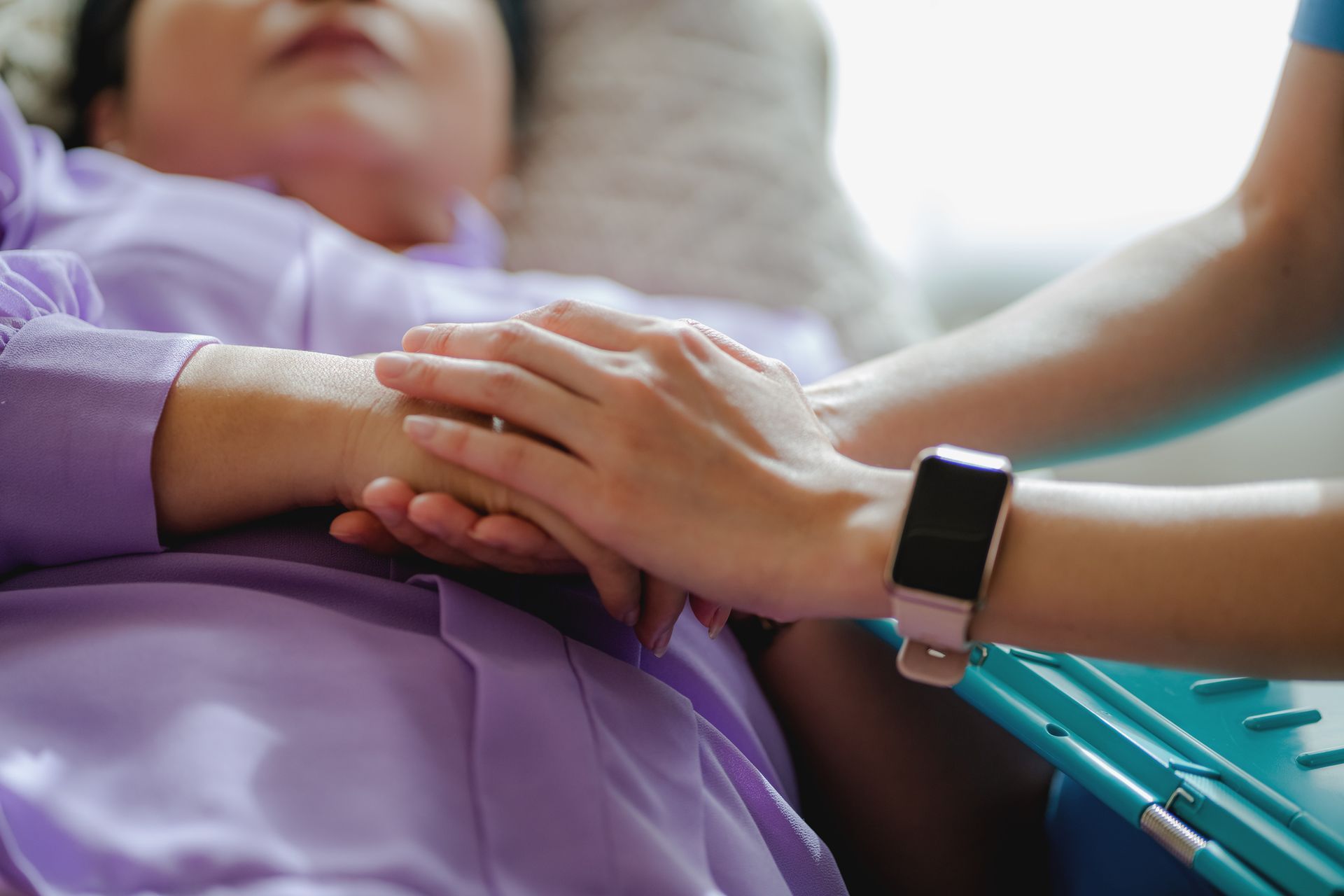 Nurse holding the hand of an elderly patient while she lies on a sofa in her home.