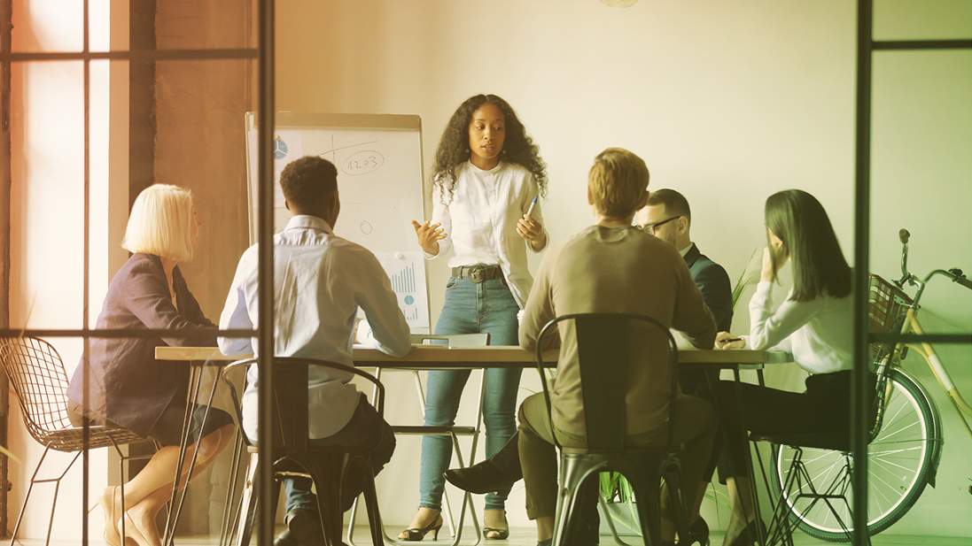 People gathered around a table in a sunlit meeting room, with a woman speaking at the front.