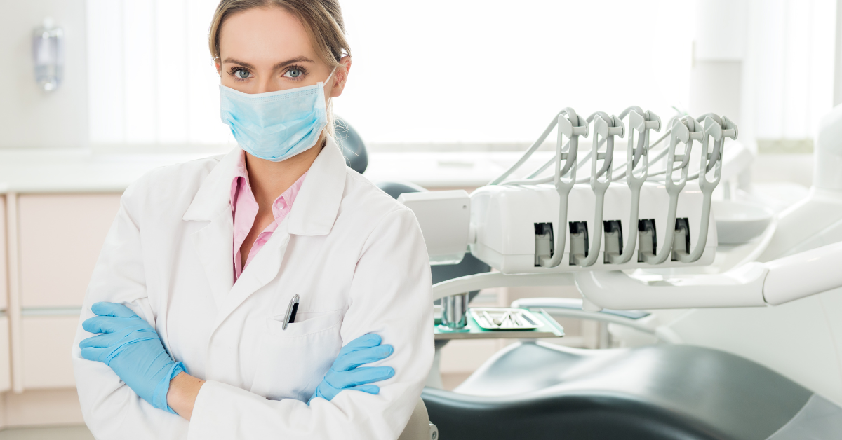 Uma dentista usando máscara e luvas azuis está em frente a uma cadeira odontológica.