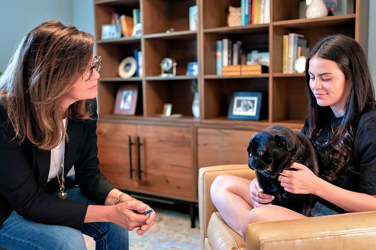 Two women seated in a living room, one holding a small black dog, with wooden shelves in the background.