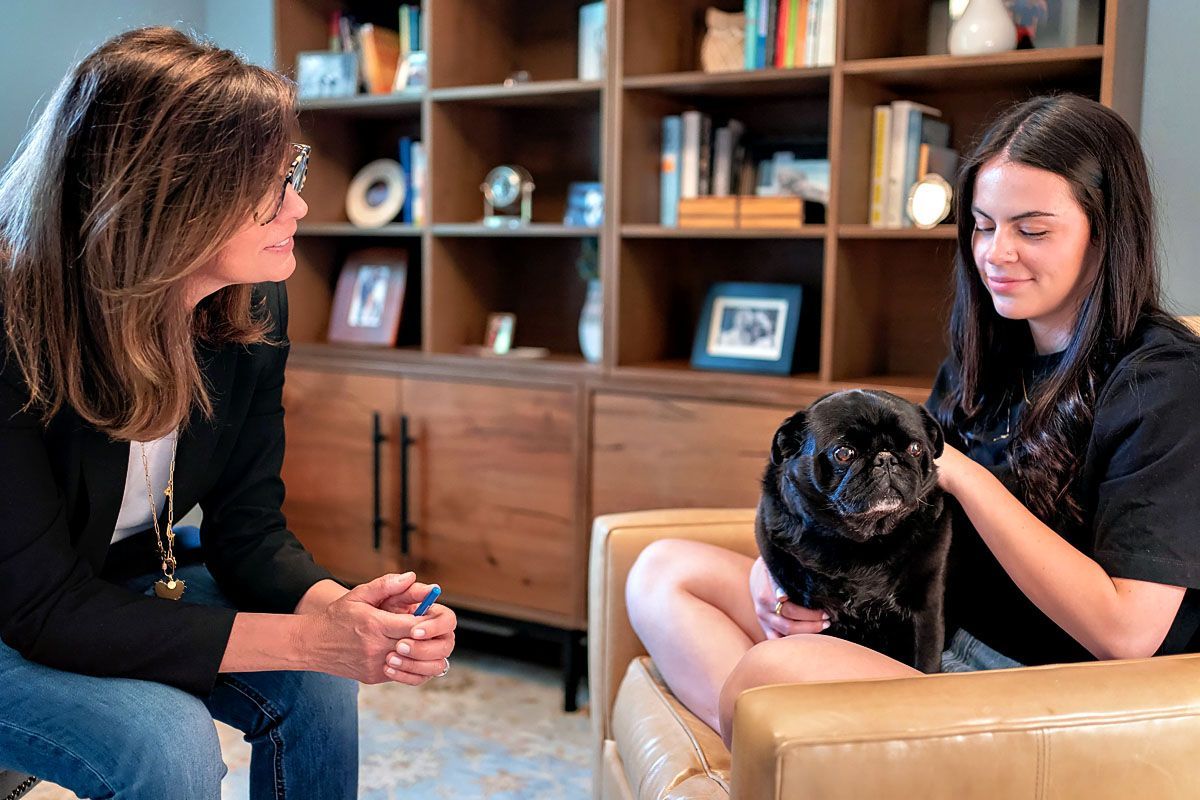 Two people sitting in the therapy office setting with a black pug on a lap, facing each other during a conversation.