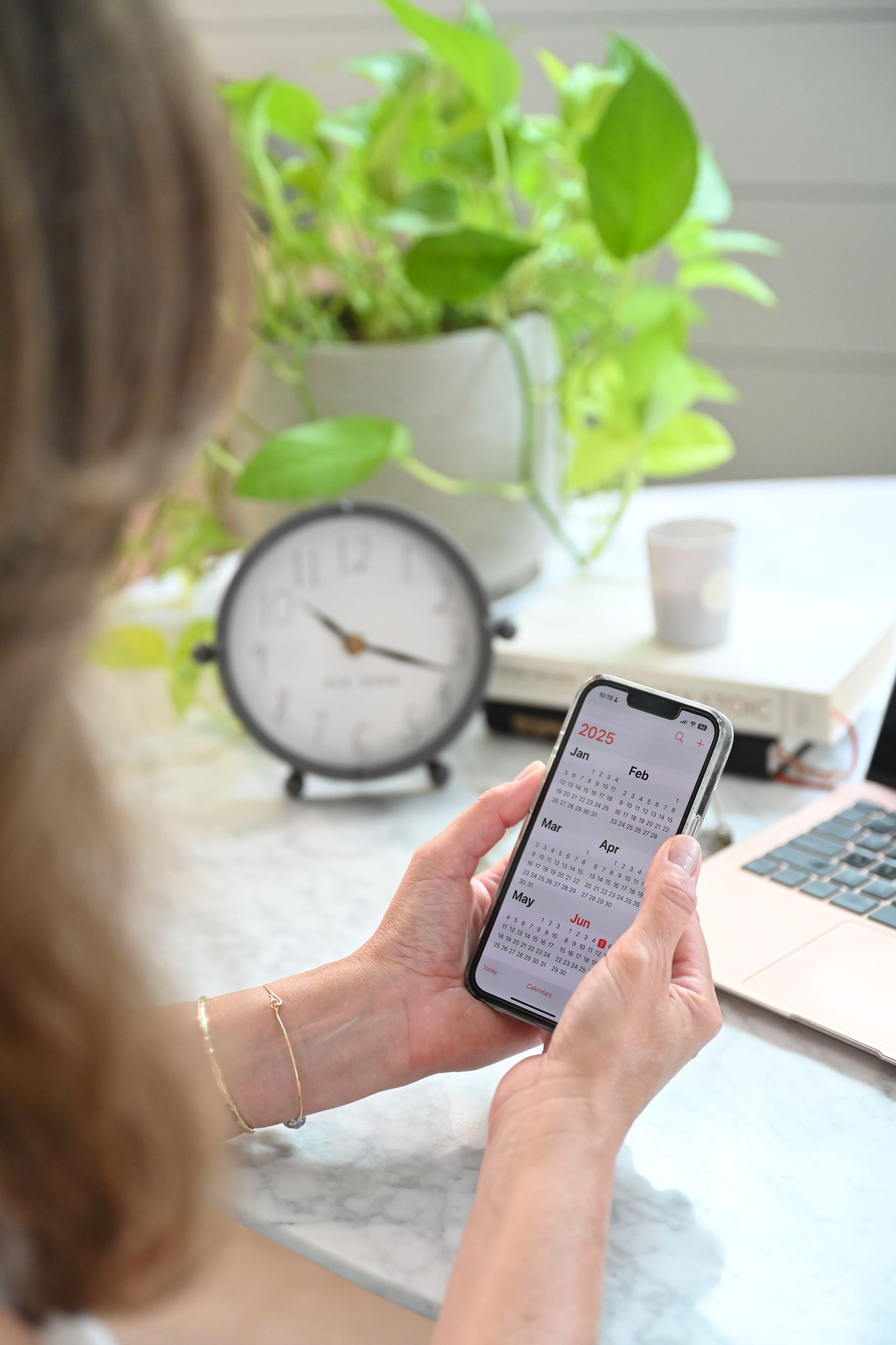 A person holds a phone displaying a calendar app at a desk with a clock, a plant, and a laptop.