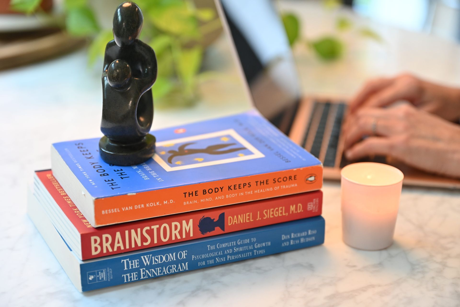 A stack of three books topped with a small sculpture sits next to a glowing candle and hands typing on a laptop.