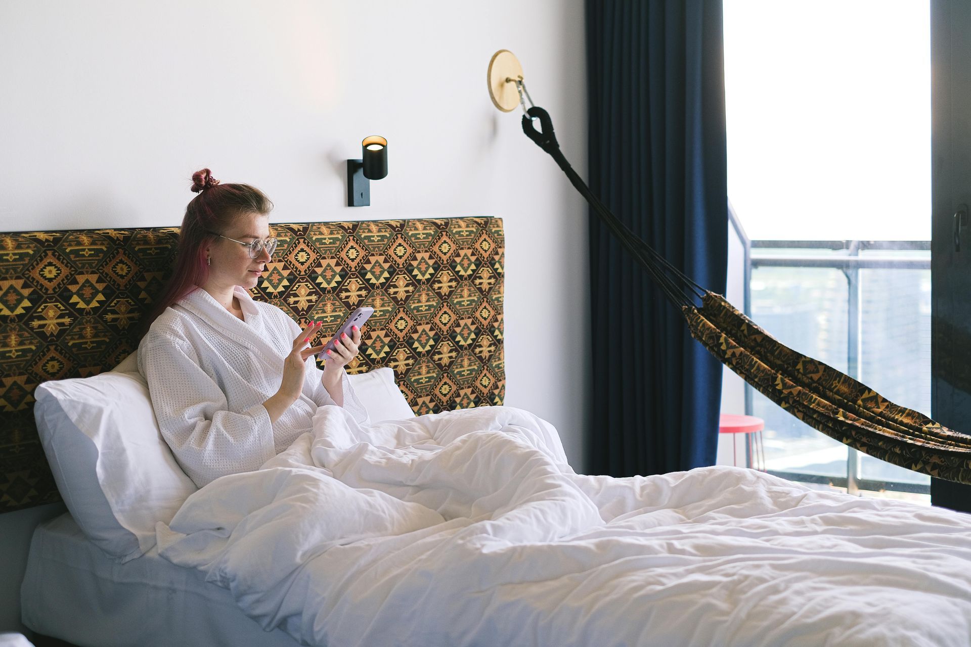 A person in a white robe sits up in a bed with a patterned headboard, looking at a smartphone with a hammock nearby.