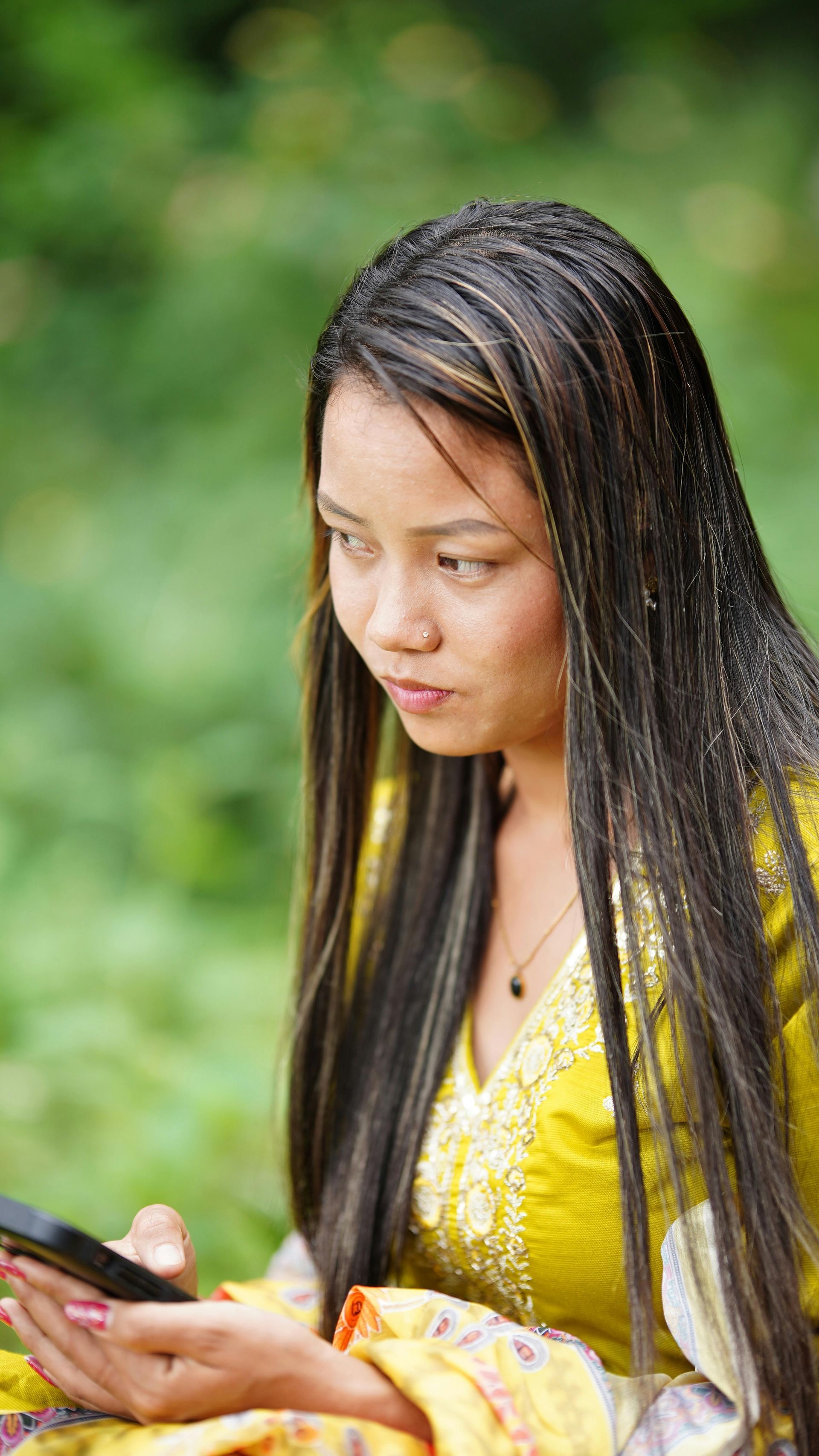 A person with long, dark, braided hair wearing a yellow patterned top looks down at a smartphone in an outdoor setting.