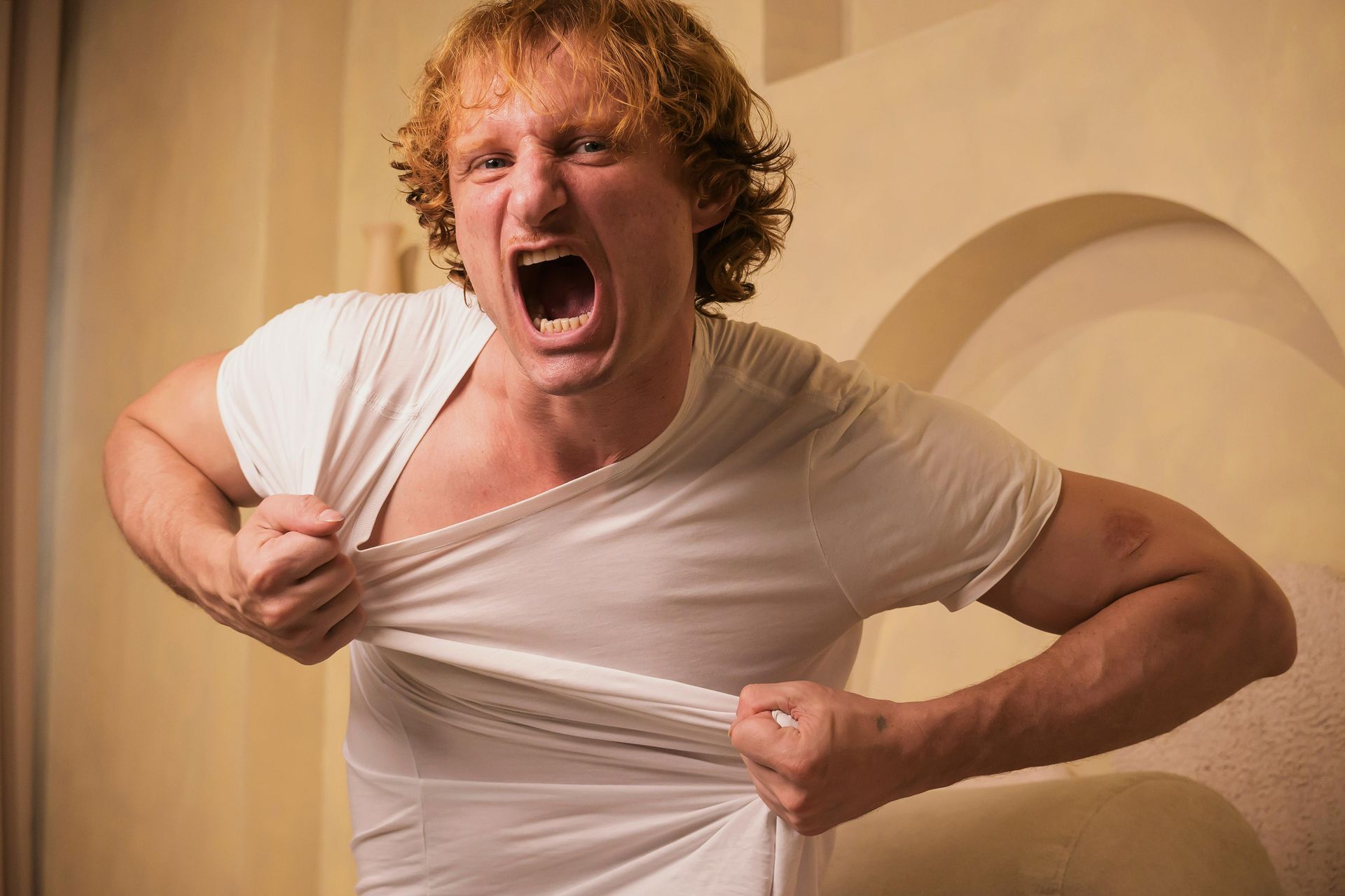A person with curly hair screams while pulling at the neckline of their white t-shirt in an indoor setting.