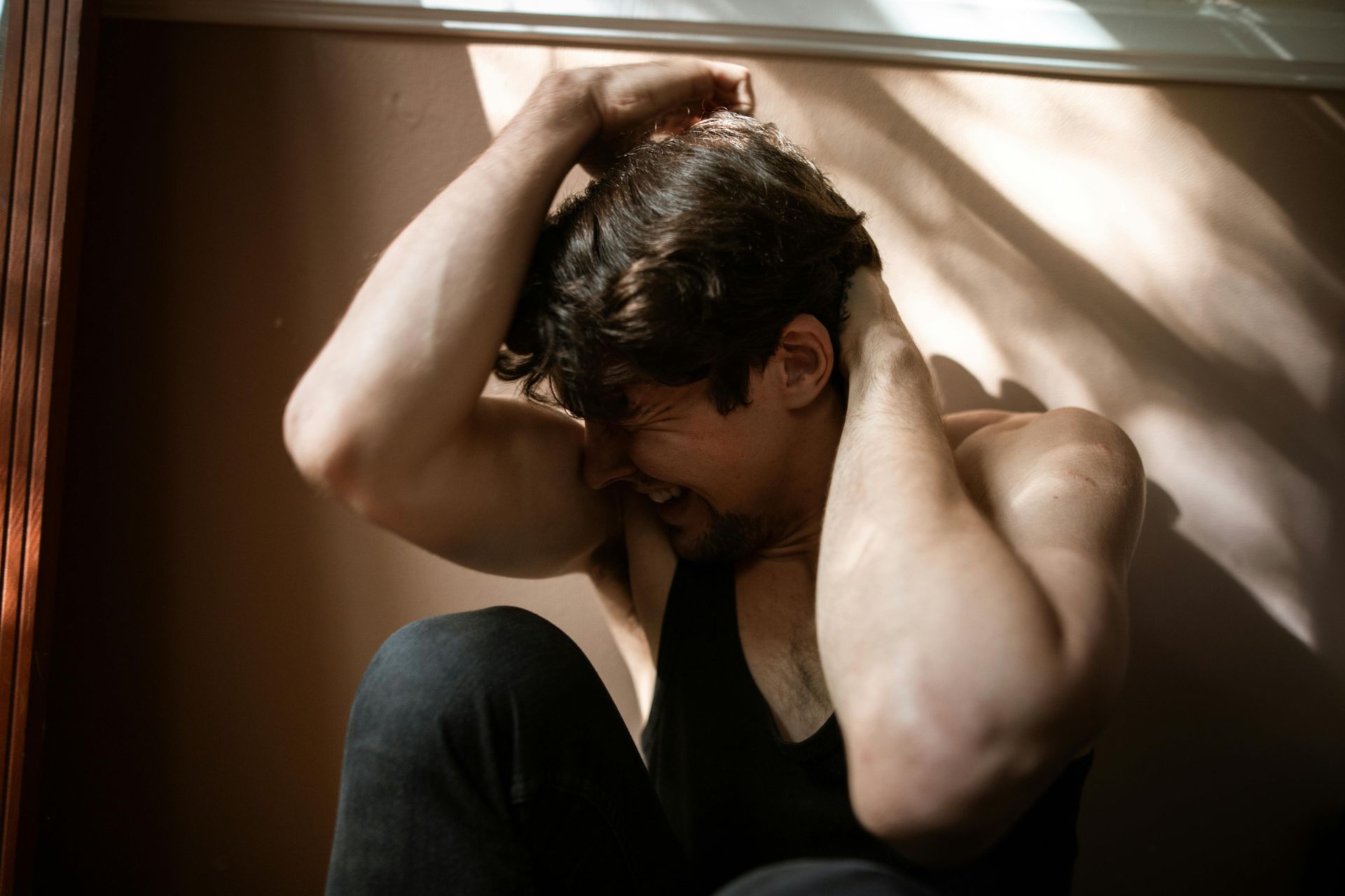 A person sitting against a wall with their hands behind their head, expressing distress with a pained facial expression.