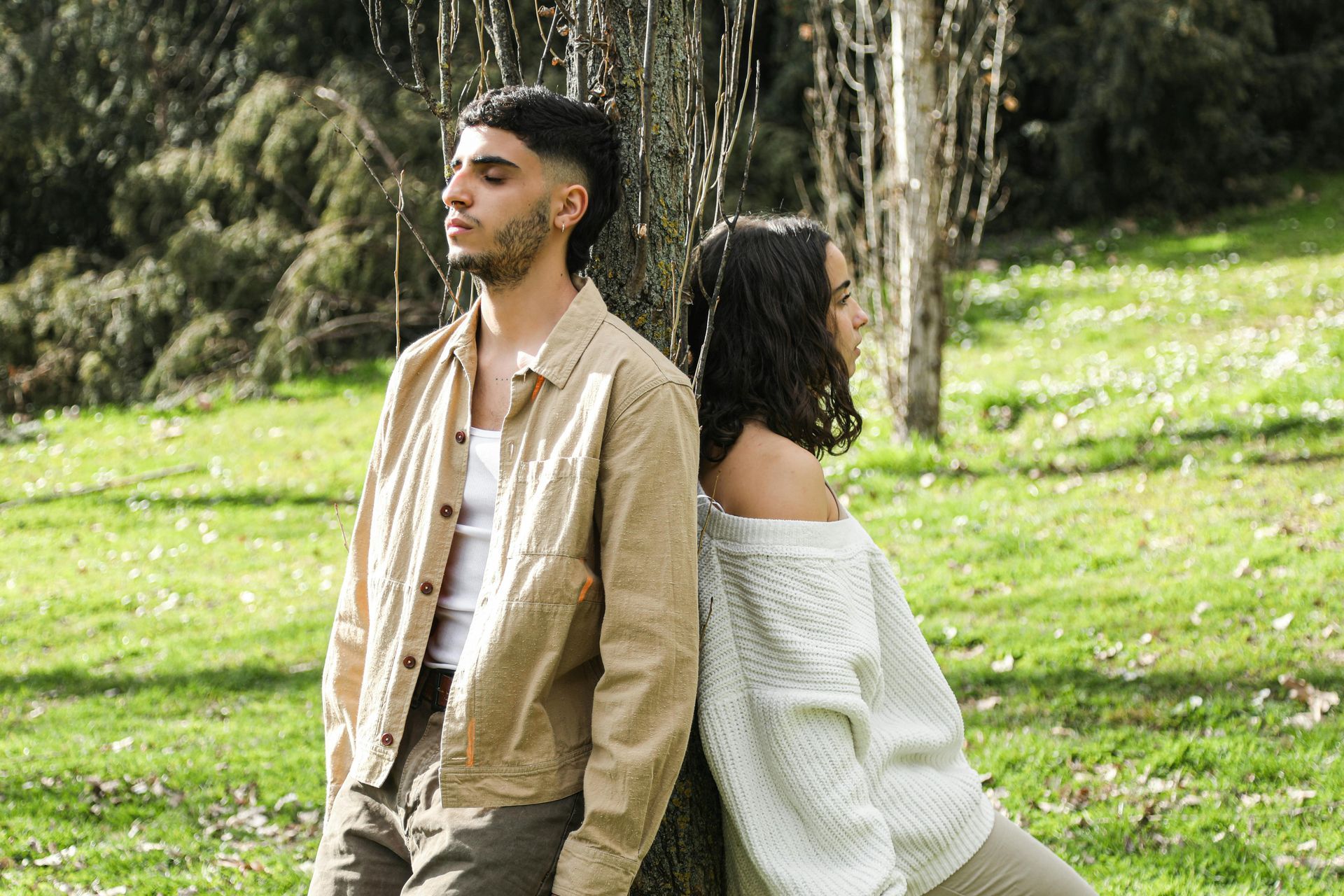 A couple standing back-to-back in a sunny park, one facing left and the other right.